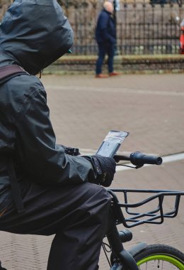 Lahey, Hollanda - 03 Ocak 2022: yağmurluk giyen bir kişi bisiklet eyerinde oturmuş akıllı bir telefonun ekranını kontrol ediyor.