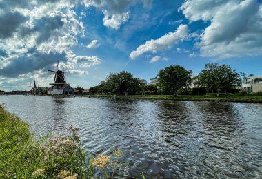 Leidschendam, Hollanda - 24 Ağustos 2021: Bulut oluşumlu bir nehir veya kanal boyunca tarihi bir rüzgar değirmeni