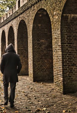 Amersfoort, Hollanda - 25 Ekim 2020: Kapüşonlu bir adam kaldırımda yürüyor güçlendirilmiş şehir surlarının kemerleri boyunca