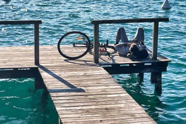 Menthon saint bernard, France - October 10 2021 : a young man with his bicycle is sunbathing and having a break on a wooden pier on the mountain lake clipart