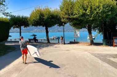 Menthon saint Bernard, France - September 12 2021 : a senior caucasian man dressed in short is carrying a surf board towards the mountain lake of annecy clipart