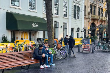 The Hague, Netherlands - December 16 2021 : two teenagers are sitting on a street bench outside eating un healthy fast food from american outlet clipart