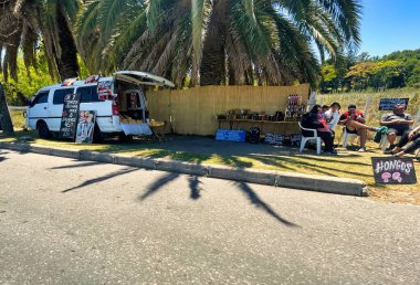 colonia del sacramento, uruguay - november 2 2022: a small stand a long the road underneath a palm tree has a table and seats and people enjoy a local fish dish clipart