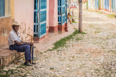 Trinidad, Küba - 15 Temmuz 2018: Trinidad 'da bir kaldırım taşı sokağında yaşlı bir adam sıcak öğleden sonra ışığında renkli binalarla çevrili oturuyor.