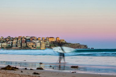 Bondi Plajı, Avustralya - 20 Ekim 2009: Bir sörfçü alacakaranlıkta Bondi Sahili 'nin kumlu kıyılarında geziniyor ve arka planda dalgalar ve kıyı evleri var..