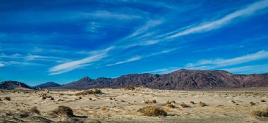 Uçsuz bucaksız kumlu arazi parlak mavi gökyüzünün altındaki görkemli dağlara zıt düşüyor..