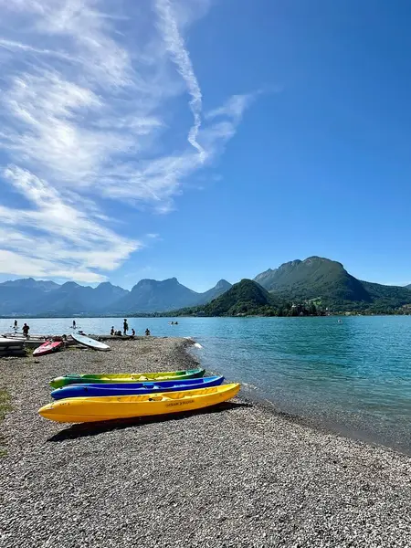Talloires, Fransa - 09 Eylül 2024: İnsanlar Annecy Gölü 'nün berrak sularının tadını çıkarırken renkli kanolar çakıllı bir plajda dinleniyor. Görkemli dağlar, parlak bir manzaranın altındaki görkemli manzarayı çerçeveler.