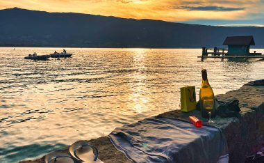 Menthon St. Bernard, Fransa - 02 Ekim 2021: Bir şarap şişesi, terlik ve havlu gün batımında dağ gölünün kıyısındaki piknik malzemeleri arasındadır.