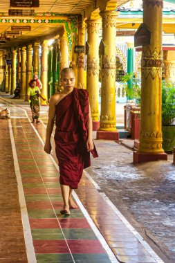 Bago, Myanmar - 3 Aralık 2012: Geleneksel giyinmiş bir keşiş hareketli bir tapınakta renkli bir patika boyunca yürür. Heykeller ve insanlar yakınlarda görünüyor..