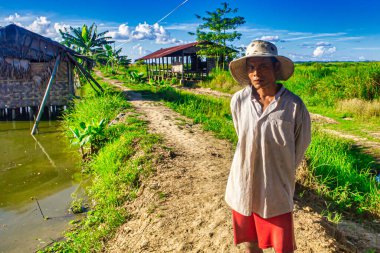 Bago, Myanmar - Aralık 03 2012: Bir adam Myanmar 'da yeşil ve geleneksel kulübelerle çevrili bir su yolunun yanında duruyor..
