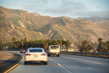 Santa Barbara, ABD - 20 Şubat 2020: Elektrikli otomobiller palmiye ağaçları ve dağlarla çevrili manzaralı bir yol boyunca sürüyorlar