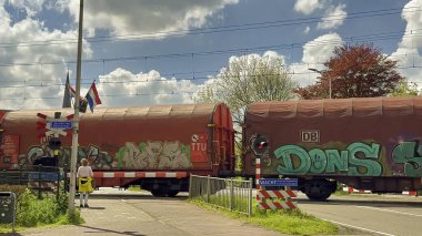 Sassenheim, Hollanda - 29 Nisan 2024: Hollanda 'da bir demiryolu geçidinde güneşli bir nisan gününde yük treninin geçişini bekleyen arabalar ve yayalar