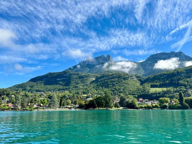 Talloires, Fransa - Eylül 06 2024: Annecy Gölü 'nün sakin suları yukarıda kabarık bulutlar sürüklenirken canlı mavi gökyüzünü yansıtır. Yeşillik ve büyüleyici evler sahili çevreleyen