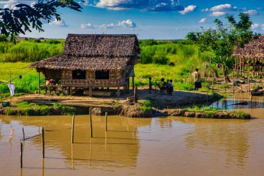 Bago, Myanmar - 03 Aralık 2012: Köylüler Myanmar kırsalındaki geleneksel sazdan bir evin önünde yemyeşil ve canlı gökyüzüyle çevrili bir şekilde dinleniyorlar..