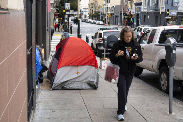 San Francisco, United States - February 13 2020 : A homeless person resides in a tent on the sidewalk in downtown San Francisco as a passerby walks by