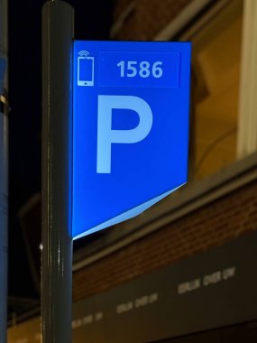 Den Haag, Hollanda - 15 Ocak 2025: 1586 sayısını gösteren parlak mavi bir park levhası gece kalabalık bir cadde ve perakende satışı yapan sürücülerin bulunduğu bölgede ayakta duruyor.