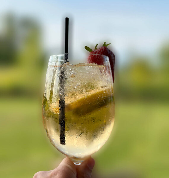 A colorful tropical cocktail garnished with a strawberry is held up showcasing refreshing ice and vibrant ingredients in a sunny outdoor setting.
