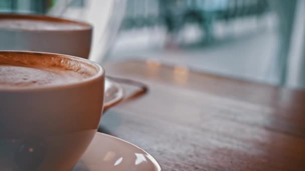 コーヒーショップのテーブルにはコーヒー2杯が置かれている 泡のあるホットカプチーノはテーブルの上で冷やされます 居心地の良いコーヒーショップでデート 白いカップからコーヒーを飲むカップル — ストック動画