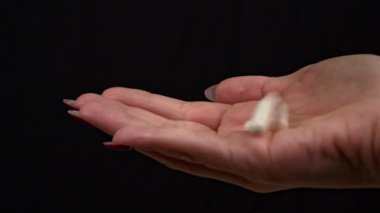 Kadın eline siyah arka plandaki bir kavanozdan beyaz haplar döküyor. Kız elinde tuttuğu kapsül dağını inceliyor. Evinde bir kadın duruyor, evinde ilaçlarla.