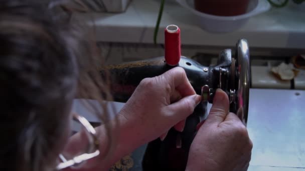 女人把缝纫线系在打字机的特写上 一个戴眼镜的老妇人在一台旧缝纫机上缝补 女裁缝把机器做的缝在红色的织物上 — 图库视频影像