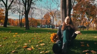 Young woman sits near a tree in an autumn park with a bouquet of maple leaves. The girl enjoys the warm autumn and basks in the sun. Walk in the autumn sunny park.
