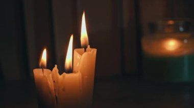 Candles burn in dark against background of a shelf with old books. Atmosphere of old library with yellow candles. Night reading of an old book in low light. Library at night, reading old literature.