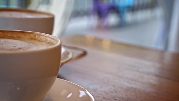 コーヒーショップのテーブルにはコーヒー2杯が置かれている 泡のあるホットカプチーノはテーブルの上で冷やされます 居心地の良いコーヒーショップでデート 白いカップからコーヒーを飲むカップル — ストック動画