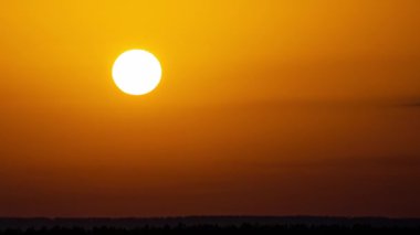 Time lapse bright orange sunset sun sets behind the clouds. The end of the day, the sun goes below the horizon. Big red hot sun in warm air distortion above the horizon.