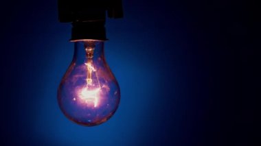 Light bulb flashes on a blue background in the dark. Slow turning on and off of a tungsten light bulb. Filament of a blinking vintage light bulb. Energy, electricity, light, life.