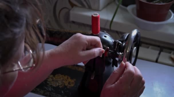 女人把缝纫线系在打字机的特写上 一个戴眼镜的老妇人在一台旧缝纫机上缝补 女裁缝把机器做的缝在红色的织物上 — 图库视频影像