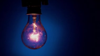 Light bulb flashes on a blue background in the dark. Slow turning on and off of a tungsten light bulb. Filament of a blinking vintage light bulb. Energy, electricity, light, life.