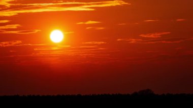 Time lapse bright orange sunset sun sets behind the clouds. The end of the day, the sun goes below the horizon. Big red hot sun in warm air distortion above the horizon.