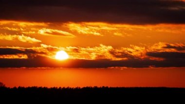 Time lapse bright orange sunset sun sets behind the clouds. The end of the day, the sun goes below the horizon. Big red hot sun in warm air distortion above the horizon.