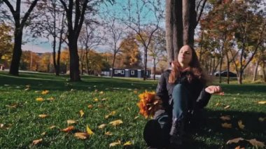 Young woman sits near a tree in an autumn park with a bouquet of maple leaves. The girl enjoys the warm autumn and basks in the sun. Walk in the autumn sunny park.