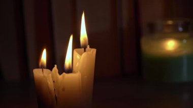 Candles burn in dark against background of a shelf with old books. Atmosphere of old library with yellow candles. Night reading of an old book in low light. Library at night, reading old literature.