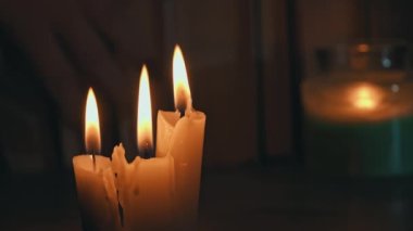 Candles burn in dark against background of a shelf with old books. Atmosphere of old library with yellow candles. Night reading of an old book in low light. Library at night, reading old literature.