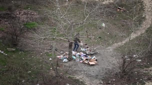 ホームレスは大量の廃棄物の隣にゴミを分別しています ホームレスの男が庭のゴミを掘っている ゴミの分別ボトルの引渡し — ストック動画