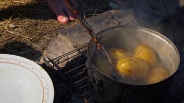 Patatesler gün boyunca açık bir ateşin üzerinde büyük bir kazanda pişirilir. Ormanda piknik. Tencere yanıyor ve su kaynıyor. Bir kamp tenceresi odun ateşinde yeni patatesler pişirir..