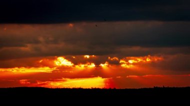 Kalkan bulutların arkasında parlak turuncu gün batımı. Güneş, günün sonunda ufkun altına iniyor. Parlayan güneşle gün batımı bulutların arasından iniyor zaman akıp gidiyor..