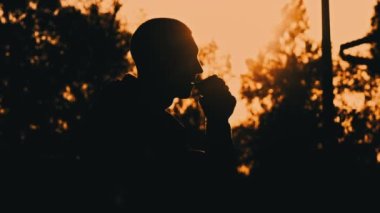 Gün batımında siluetli adam bankta kahve içiyor. Doğada akşam mis kokulu kahve. Bir adam gün batımında kahveyi sever. Doğayla baş başa düşünceler. Serbest düşünce konsepti.