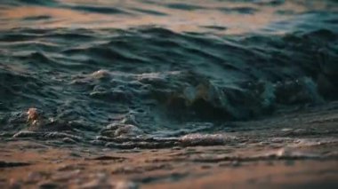 Dalgalar kıyıda günbatımını yavaş çekimde yansıtıyor. Güneşli bir yaz gününde suda dalgalanmalar. Güneş ışınlarının suya yansıması. Batan güneş görkemli dalgalara yansıyor..