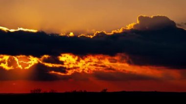 Kalkan bulutların arkasında parlak turuncu gün batımı. Güneş, günün sonunda ufkun altına iniyor. Parlayan güneşle gün batımı bulutların arasından iniyor zaman akıp gidiyor..