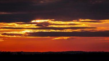 Kalkan bulutların arkasında parlak turuncu gün batımı. Güneş, günün sonunda ufkun altına iniyor. Parlayan güneşle gün batımı bulutların arasından iniyor zaman akıp gidiyor..