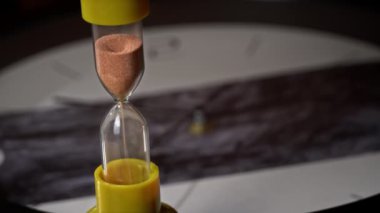 Kum saati arka plandaki sıradan mekanik saate doluyor. Kum yağıyor ve zamanı sayıyor. Zaman akıp gidiyor, zamanın geçişi. Geri sayım, dakika, saniye, saat.