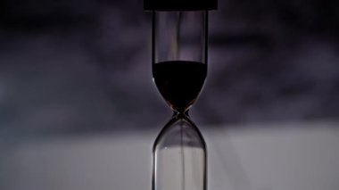 Kum saatinin silueti sıradan mekanik saatin arka planına doğru akar. Kum düşer ve zamanı sayar. Zaman akıp gidiyor, zamanın geçişi. Geri sayım, dakika, saniye, saat.