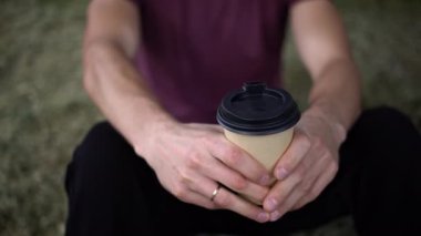 Bir adam, elinde sıcak bir kahve fincanı tutarak rahatça dışarıda oturuyor. Arka plandaki yumuşak odak, doğanın sükuneti içinde sakin bir keyif anını vurgular..