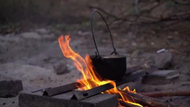 Kamp ateşinin üzerinde siyah bir tencere asılı duruyor. Tencerenin bir kabı var ve kaynar suyla dolu. Alevler görünüyor..