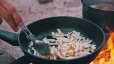 Kamp ateşinin üstünde doğranmış soğanları siyah bir tavada karıştıran bir elin yakın çekimi. Soğanlar altın sarısı ve alevler görülebilir. Çerçevenin sağında siyah bir tencere var..
