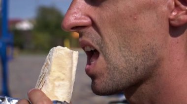 Güneşli bir günde shawarma yiyen bir adamın yakın çekimi. Bir parça ısırıyor, arkasında peynir bırakıyor..