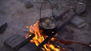 Siyah bir tencere kamp ateşinin üzerinde asılı, patatesli kaynar su. Tencerenin kablo kolu var. Alevler görünüyor..